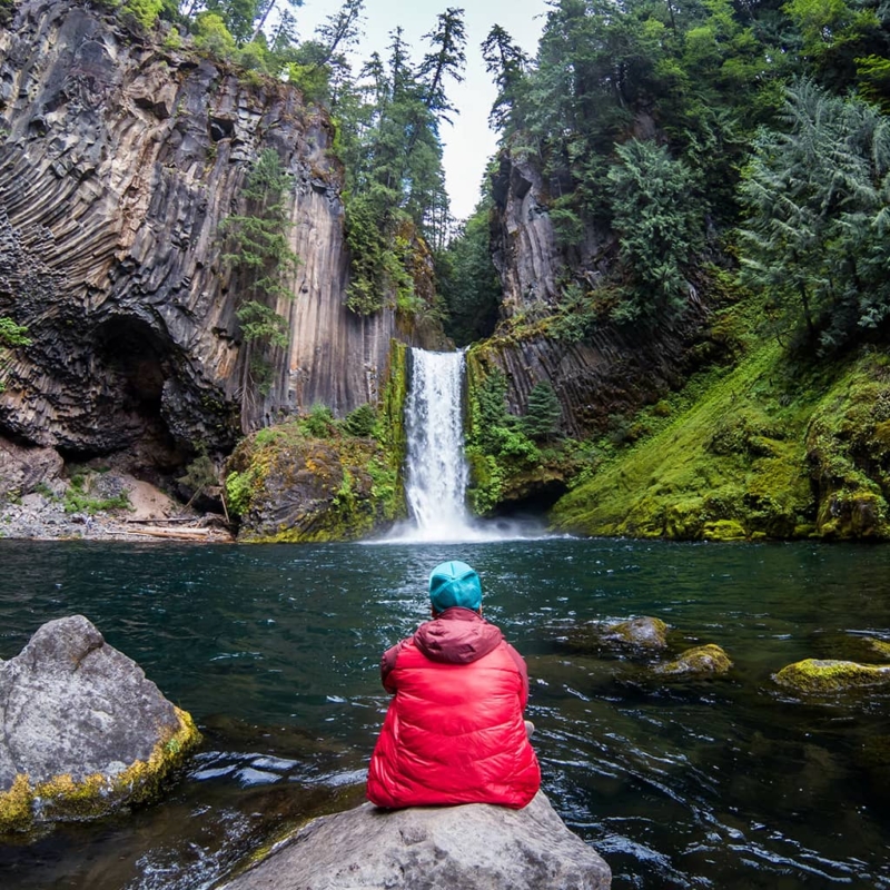 A person wearing a bright red jacket sits on a rock facing Toketee waterfall, which flows over naturally formed volcanic rocks and is surrounded by pine trees on the Umpqua River in Oregon